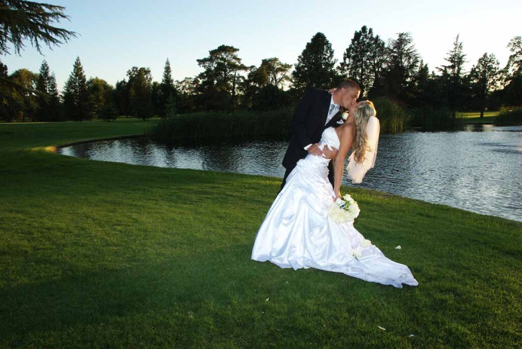Groom leans his bride back to kiss her while posing in front of a lake.