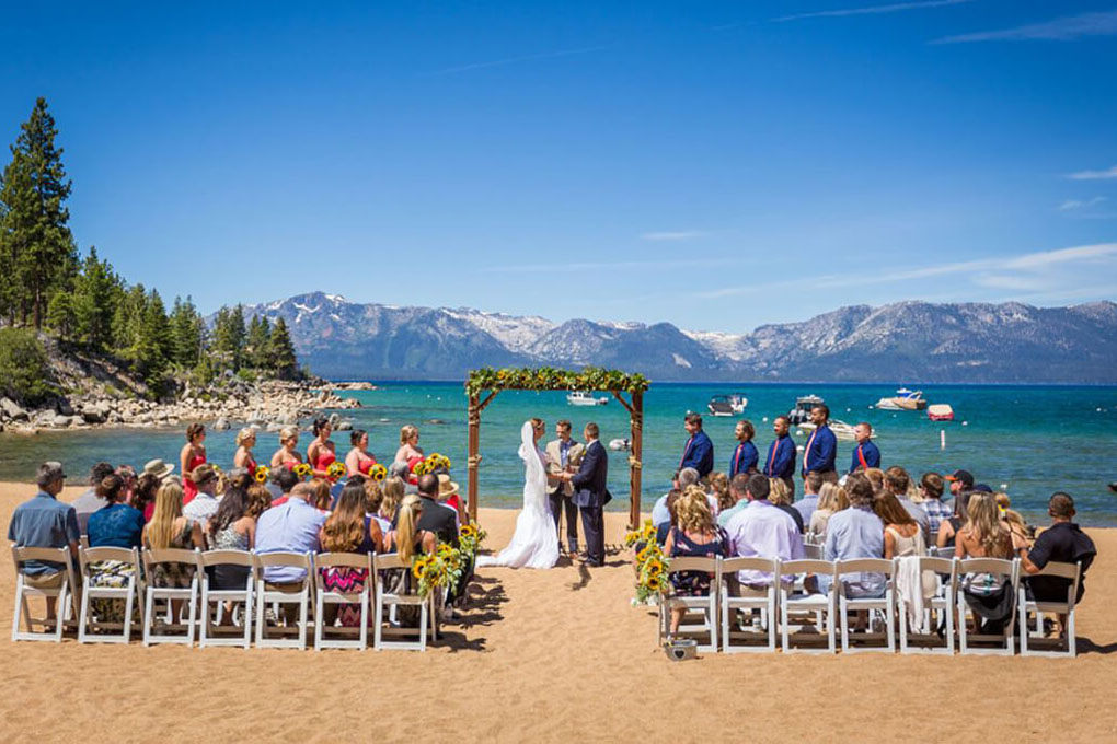 Onlooking guests during a ceremony in progress at the edge of the water under an arch.