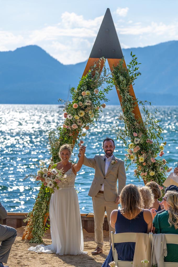 Just married twosome raise their joined hands high in front of the triangular arch decorated with flowers.