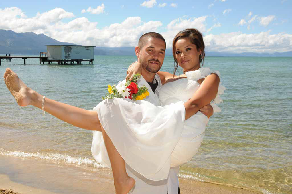 Groom carries his bride in his arms while on the beach.