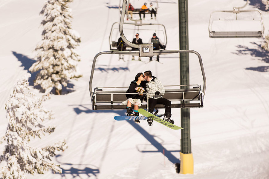 The about to be married couple with snowboards are riding up the ski lift.
