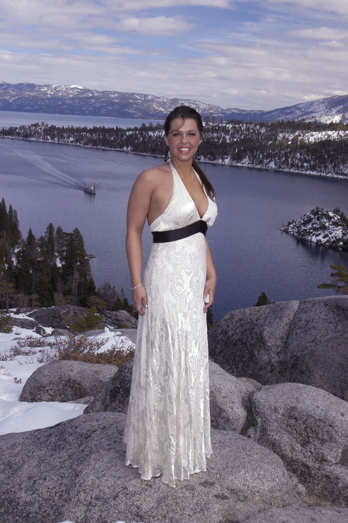 Tall formally dressed bride stands on a rock overlooking the bay.