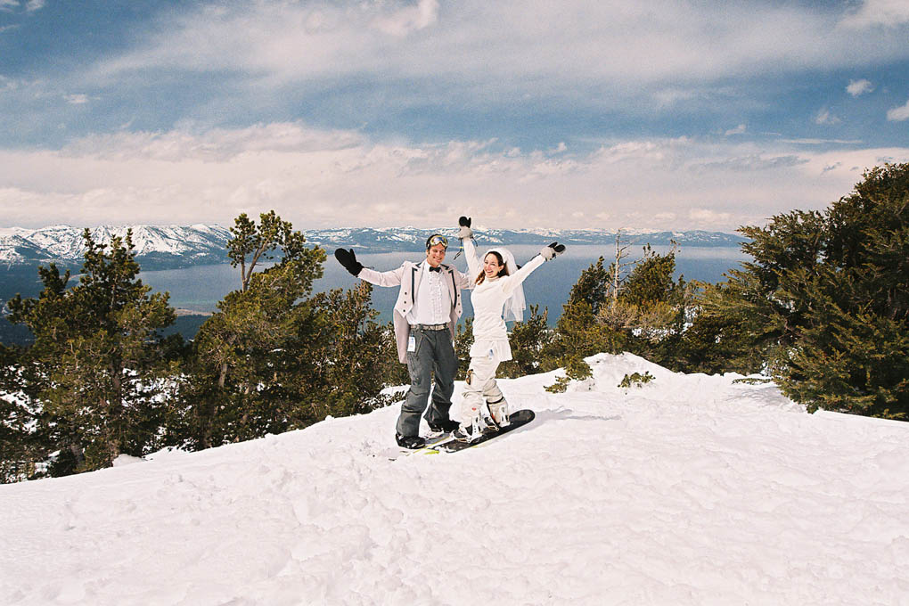 Married pair on skis raise their hands in the air on Heavenly mountaintop.