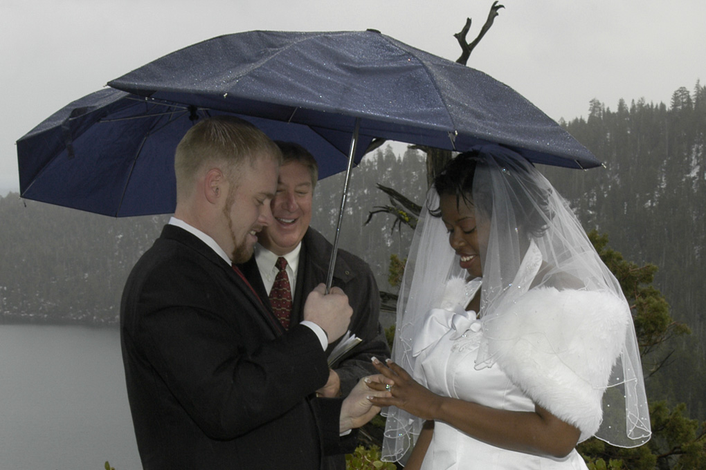 Groom slides wedding ring on bride's hand while holding an umbrella.