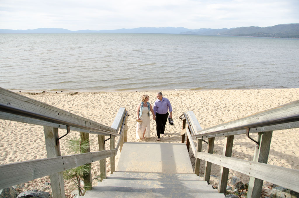 The wedded pair carrying their shoes and walking barefoot on the beach near the staircase.