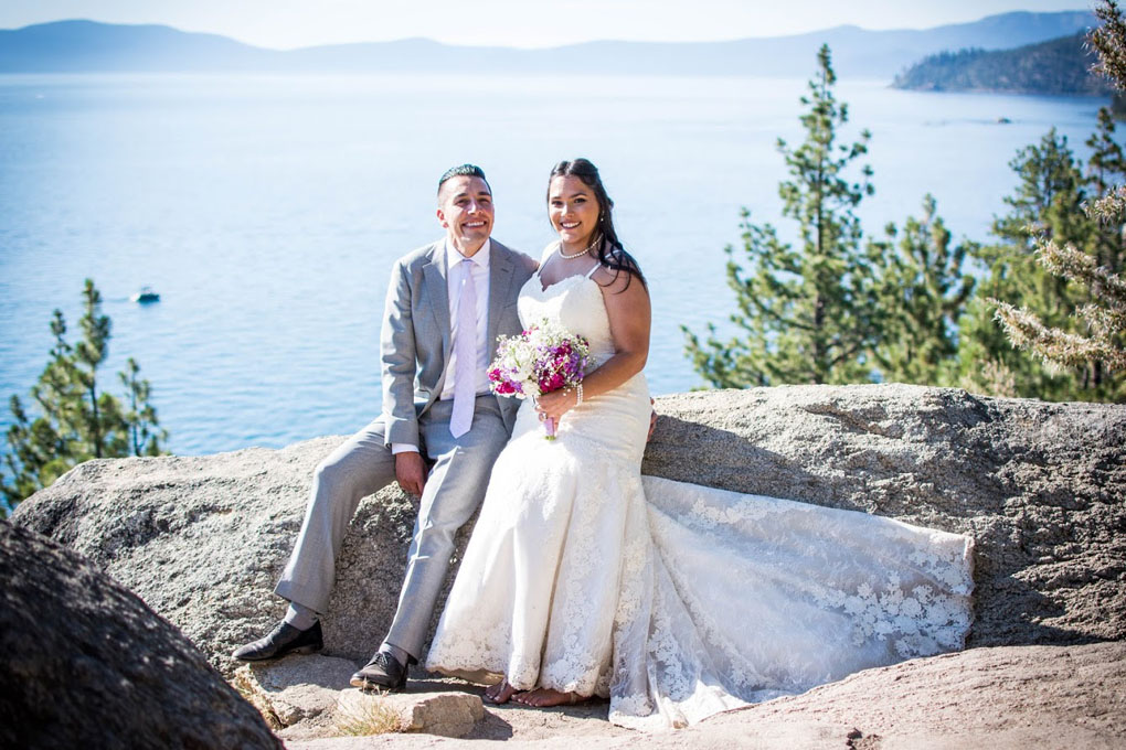 Newlyweds in formal attire sitting on a rock.